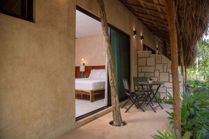 Un dormitorio con cama, mesita de noche y ventana. Hay una pequeña mesa y una silla fuera de la habitación.
