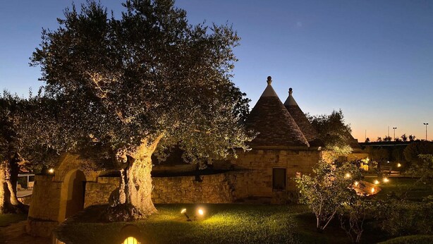 A stone building with a conical roof, surrounded by well-maintained grass and trees, illuminated by ground lights. - Relais Il Palmento (Locorotondo)