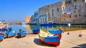 A harbor with several small boats docked, a large historic building with arched windows, and a clear blue sky. - Relais Il Palmento (Locorotondo)