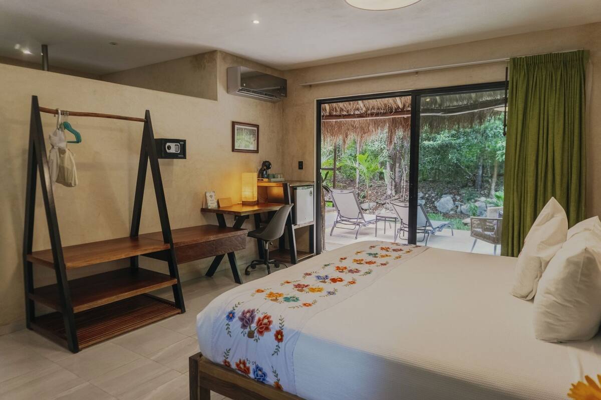 A hotel room with a bed, desk, chair, and a balcony with a view of greenery.