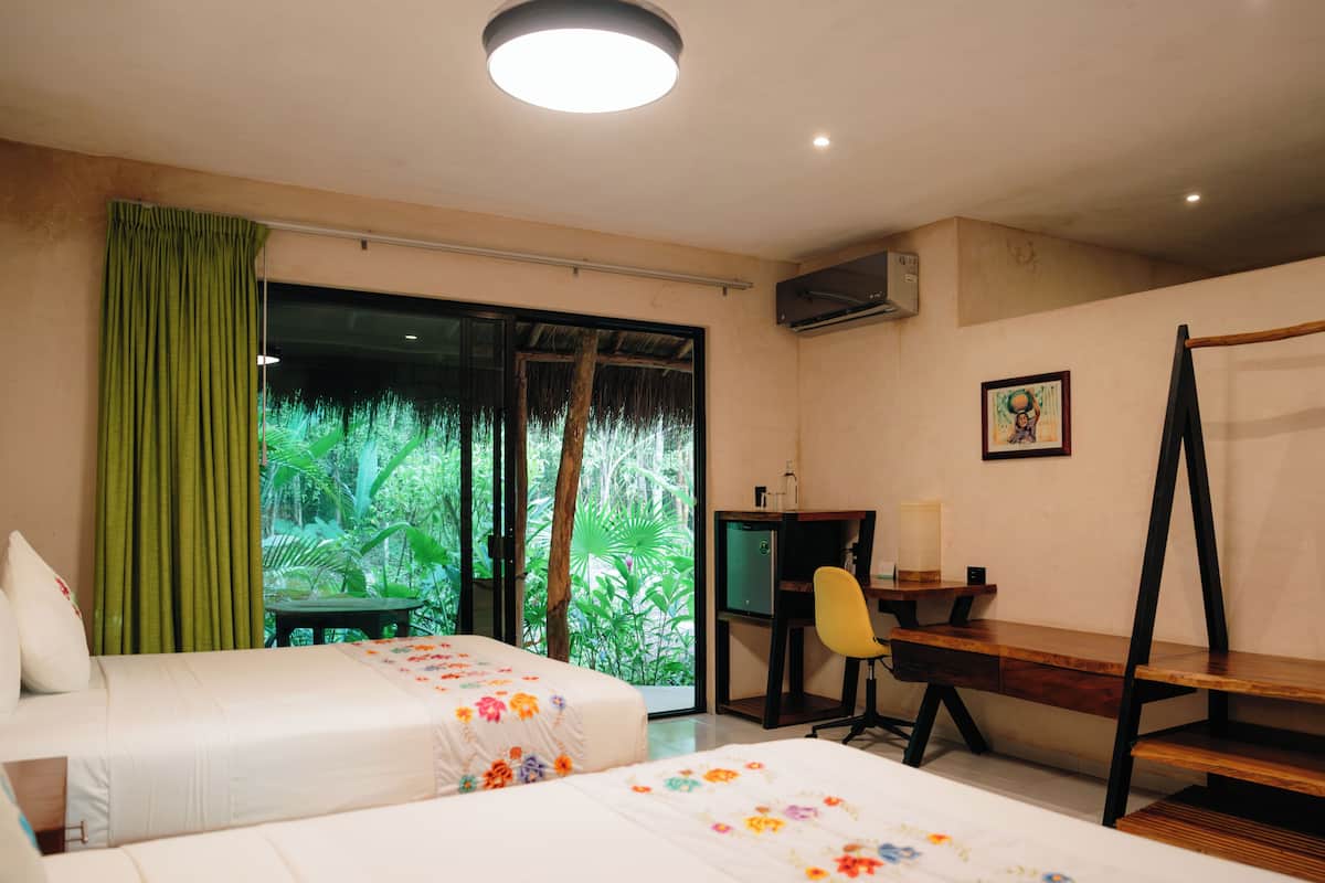 A hotel room with two beds, a desk, a chair, and a large window with a view of greenery.
