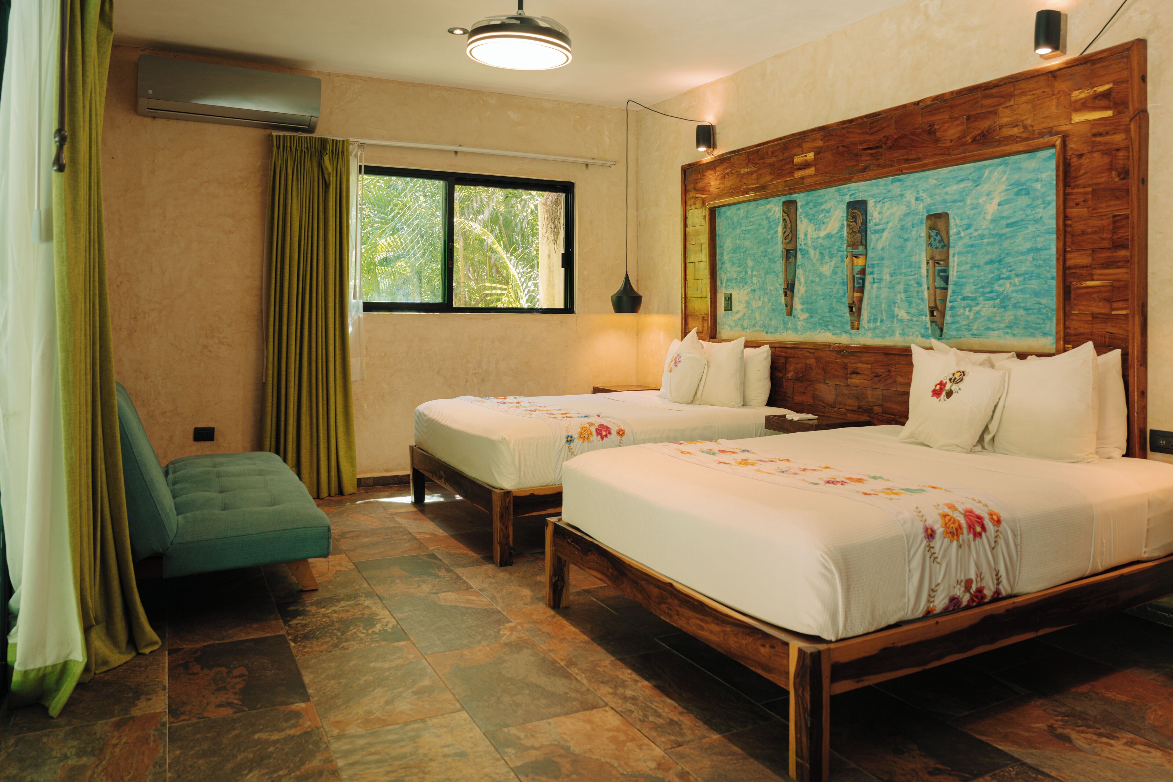 A hotel room with two single beds, a wooden headboard, a large window with curtains, a green sofa, and a wall-mounted air conditioner.