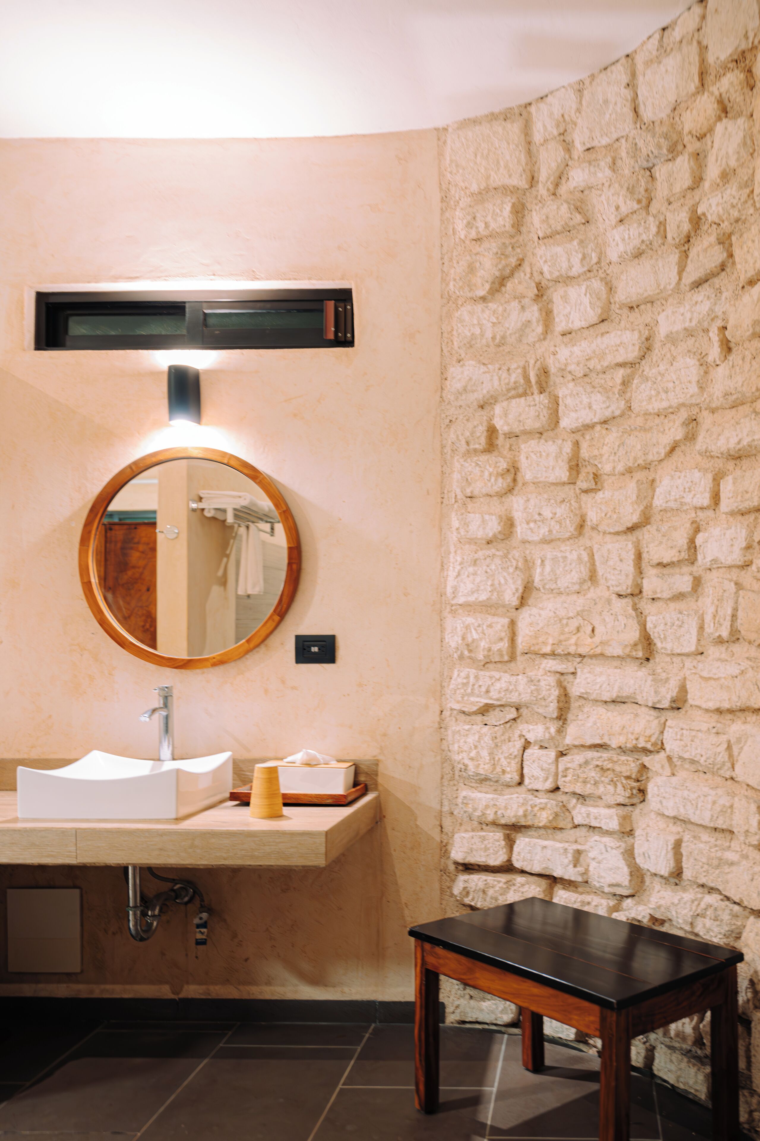 A bathroom with a stone wall, a round mirror, a sink, and a small wooden table.