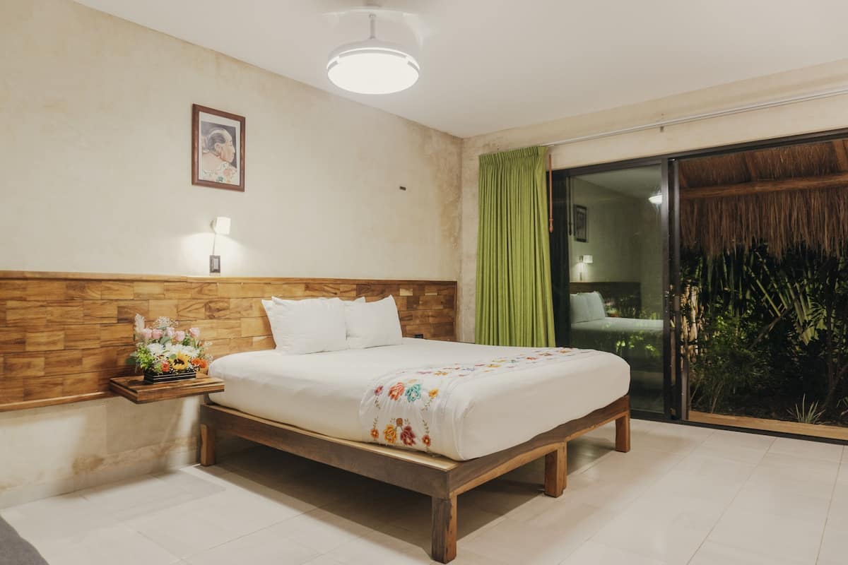 A bedroom with a wooden headboard, a bed with a floral bedspread, a nightstand with a flower arrangement, and a view of a tropical garden through a sliding door.