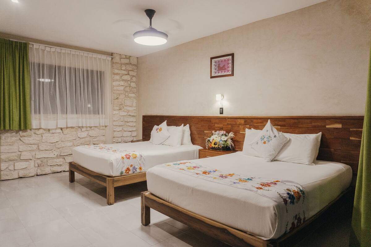 A hotel room with two beds, a stone wall, a wooden headboard, a nightstand with a vase, and a ceiling fan.