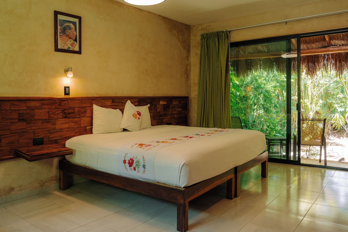 A hotel room with a wooden headboard, a large bed with floral bedding, a nightstand, a wall-mounted light, and a view of greenery through a sliding glass door.