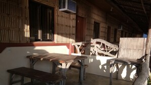 Un espace extérieur aménagé dans un style rustique, avec une table et des bancs en bois, entouré de murs en bambou et surmonté d’une structure de toit en bois.
