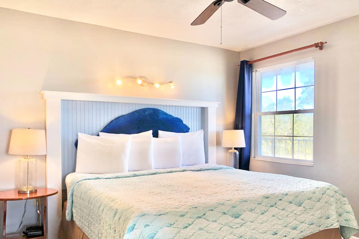 A neatly made bed with a blue headboard, two bedside lamps, and a window with curtains.
