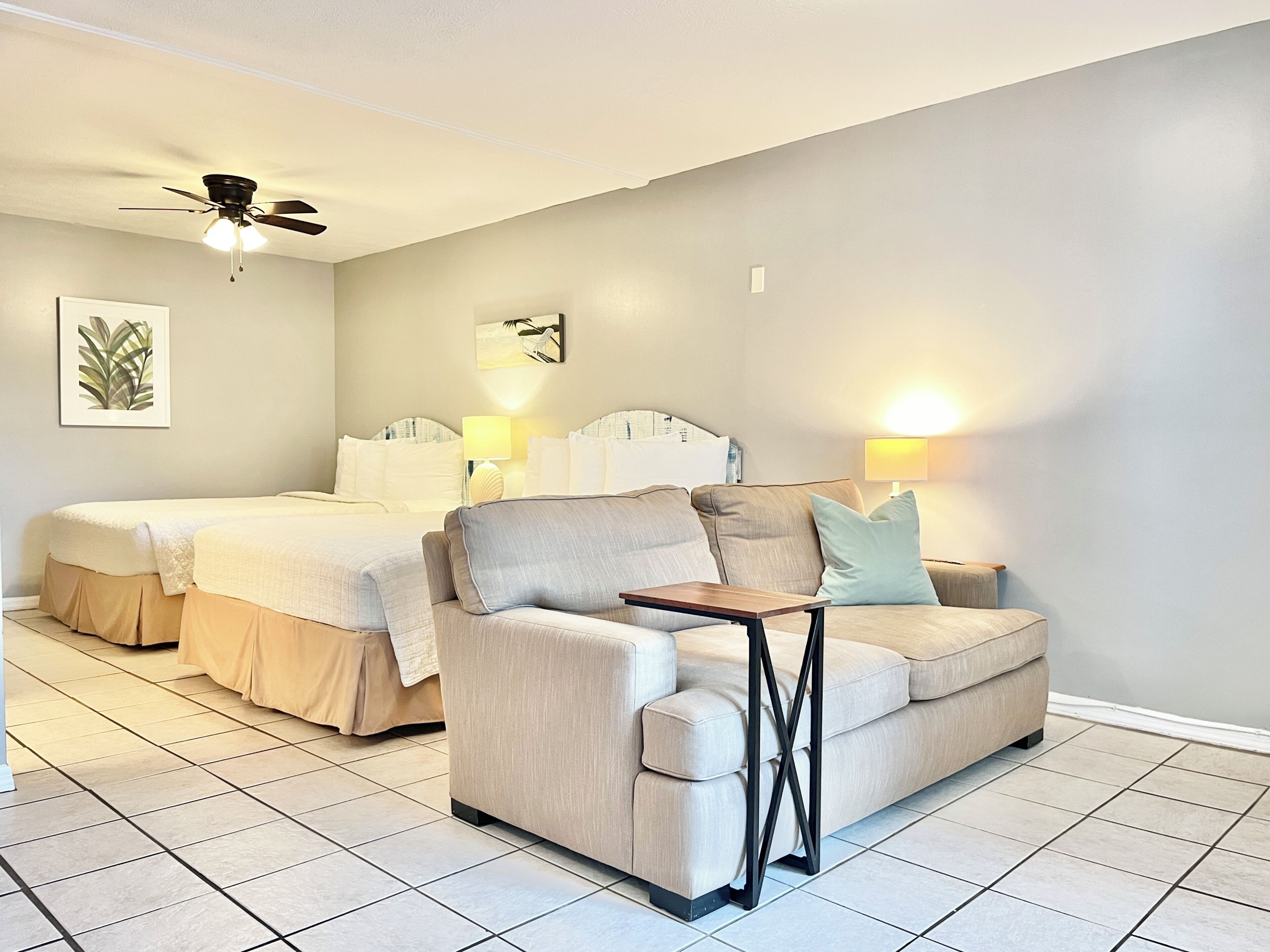 A hotel room with two beds, a sofa, a ceiling fan, and two wall-mounted lamps.