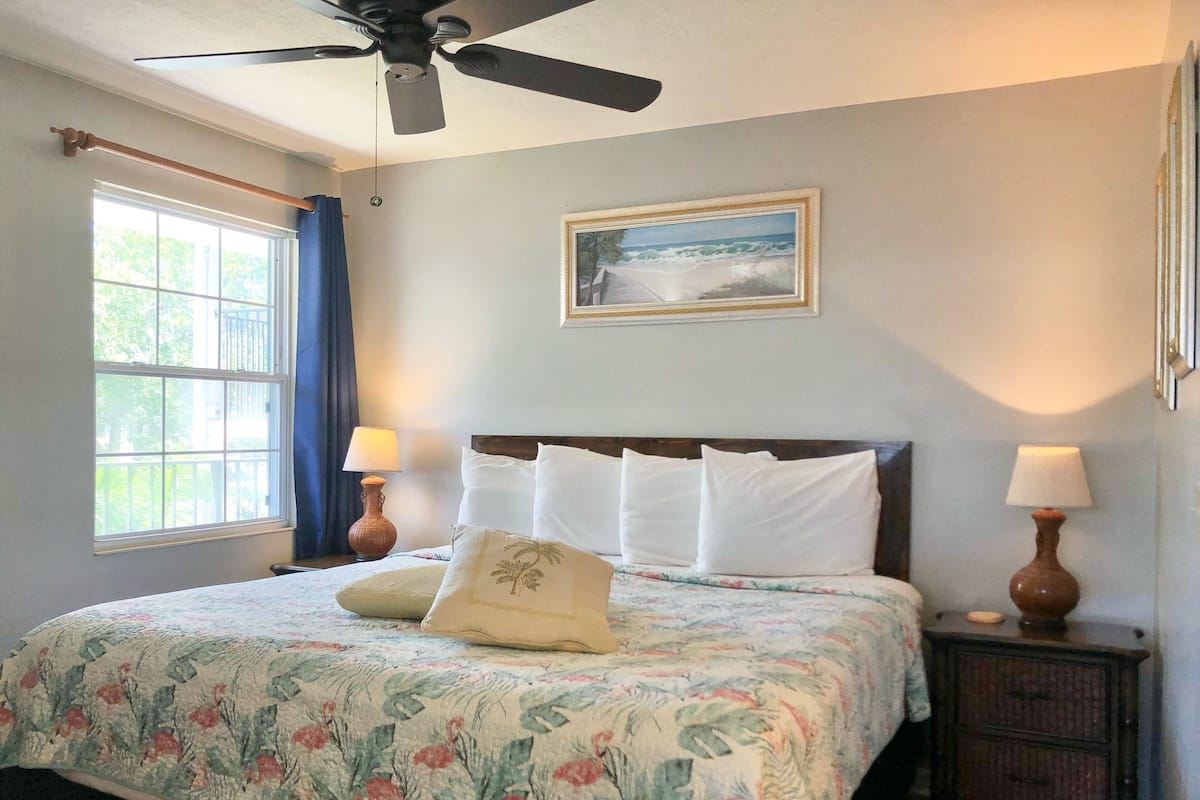 A bedroom with a bed, two bedside lamps, a ceiling fan, a window with curtains, and a framed picture on the wall.