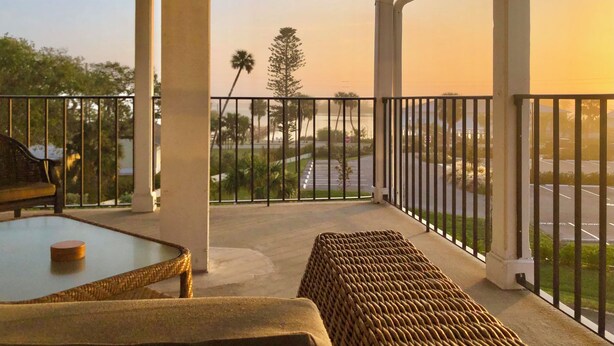 A covered balcony with a glass table, wicker furniture, and a view of a parking lot and palm trees. - Davis House Inn (Sebastian)