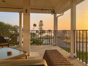 A covered balcony with a glass table, wicker furniture, and a view of a parking lot and palm trees.