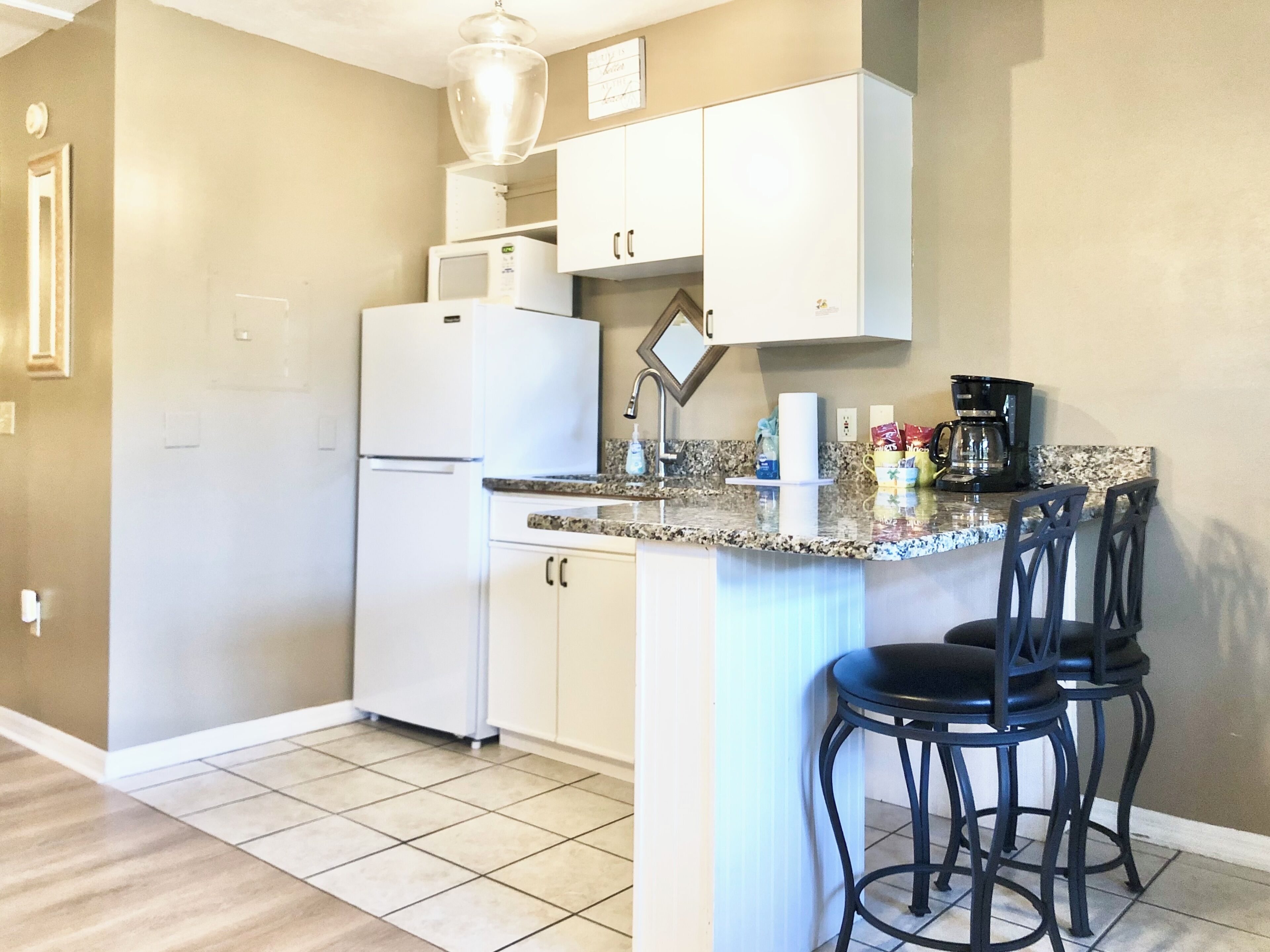 A kitchen with white cabinets, a refrigerator, a microwave, a coffee maker, and a bar stool.