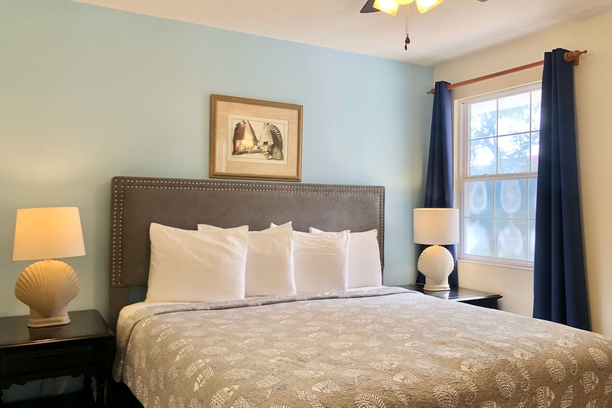 A neatly made bed with a patterned bedspread, a framed picture above it, and a window with curtains to the right.