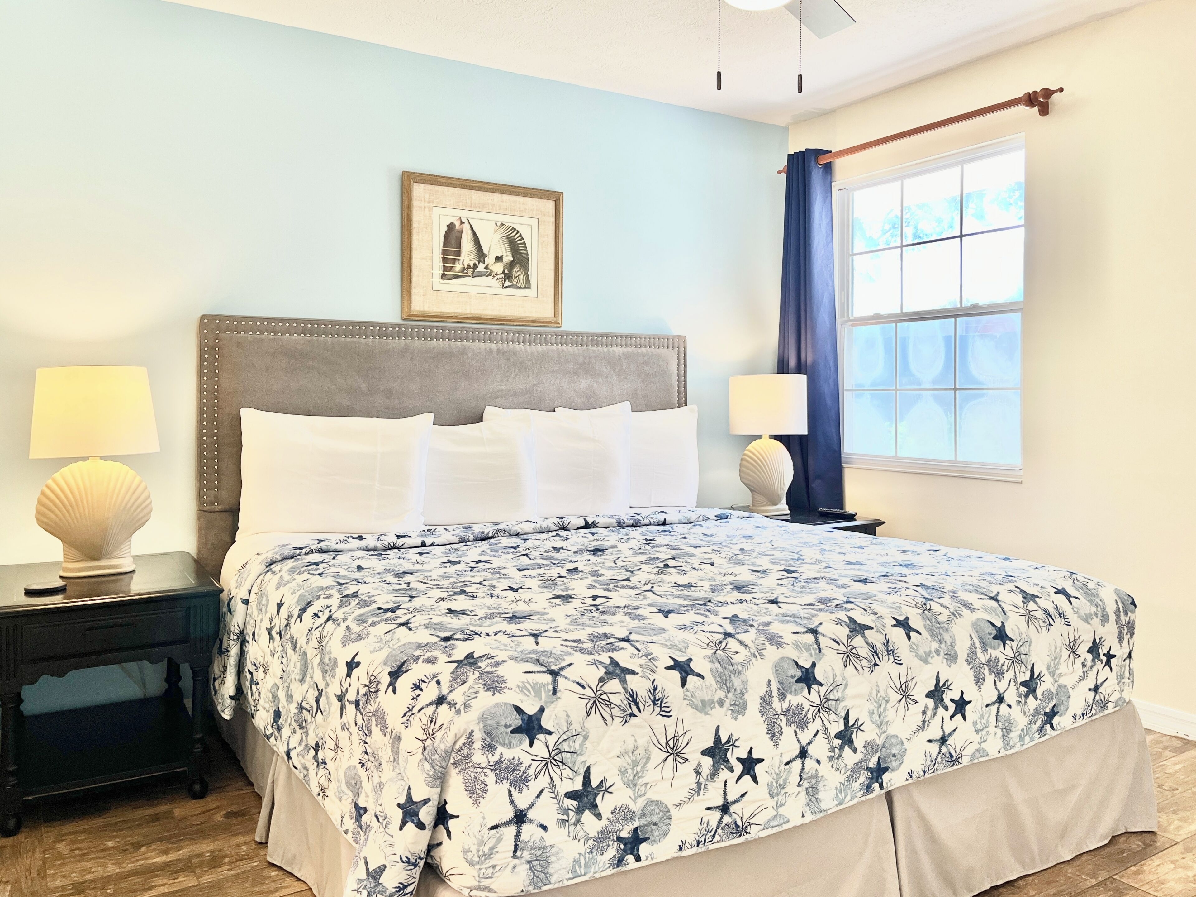 A bedroom with a bed, two nightstands, a framed picture on the wall, a window with curtains, and a ceiling fan.