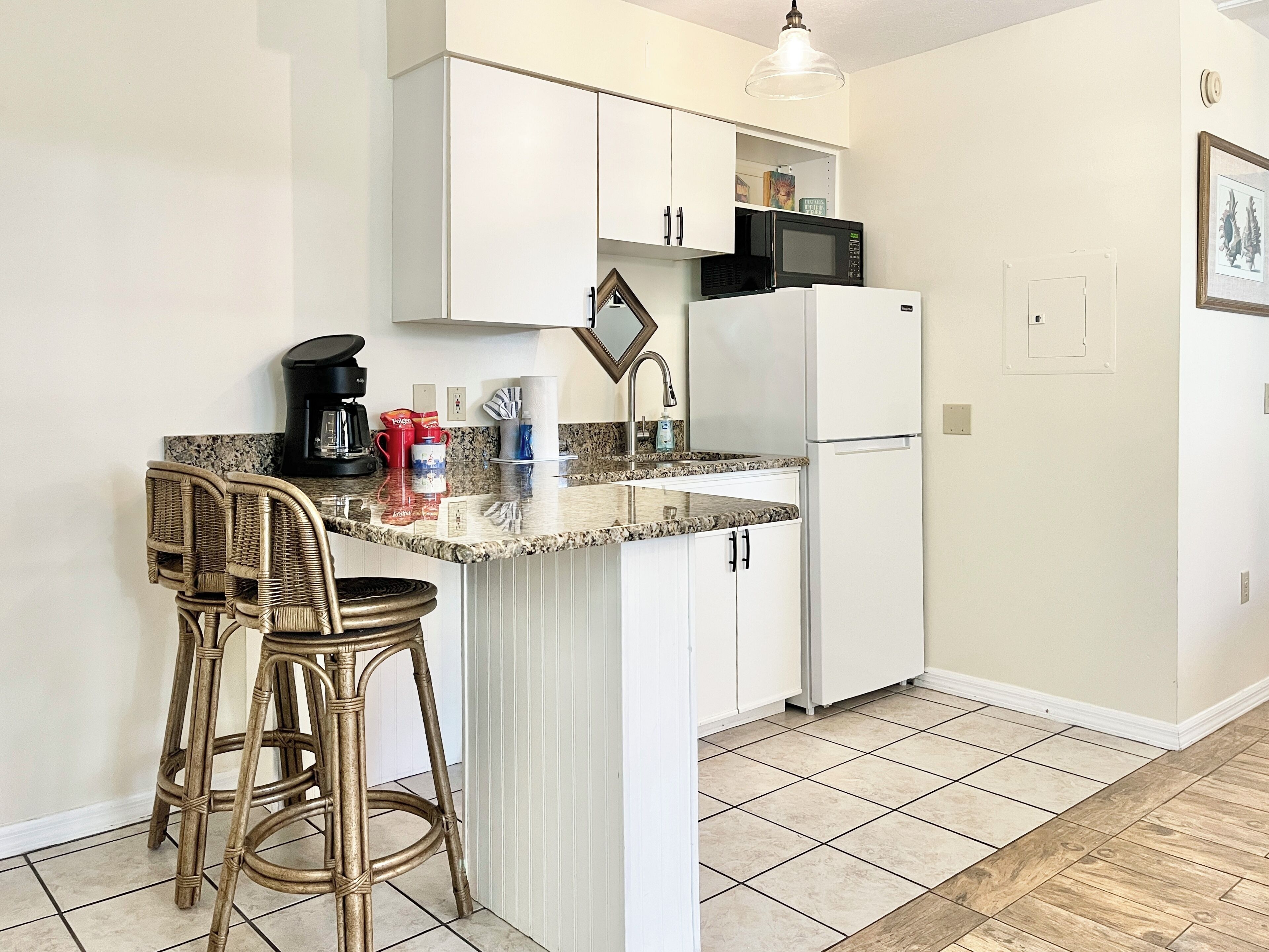 A kitchen with white cabinets, a microwave, a refrigerator, a coffee maker, and two wicker bar stools.