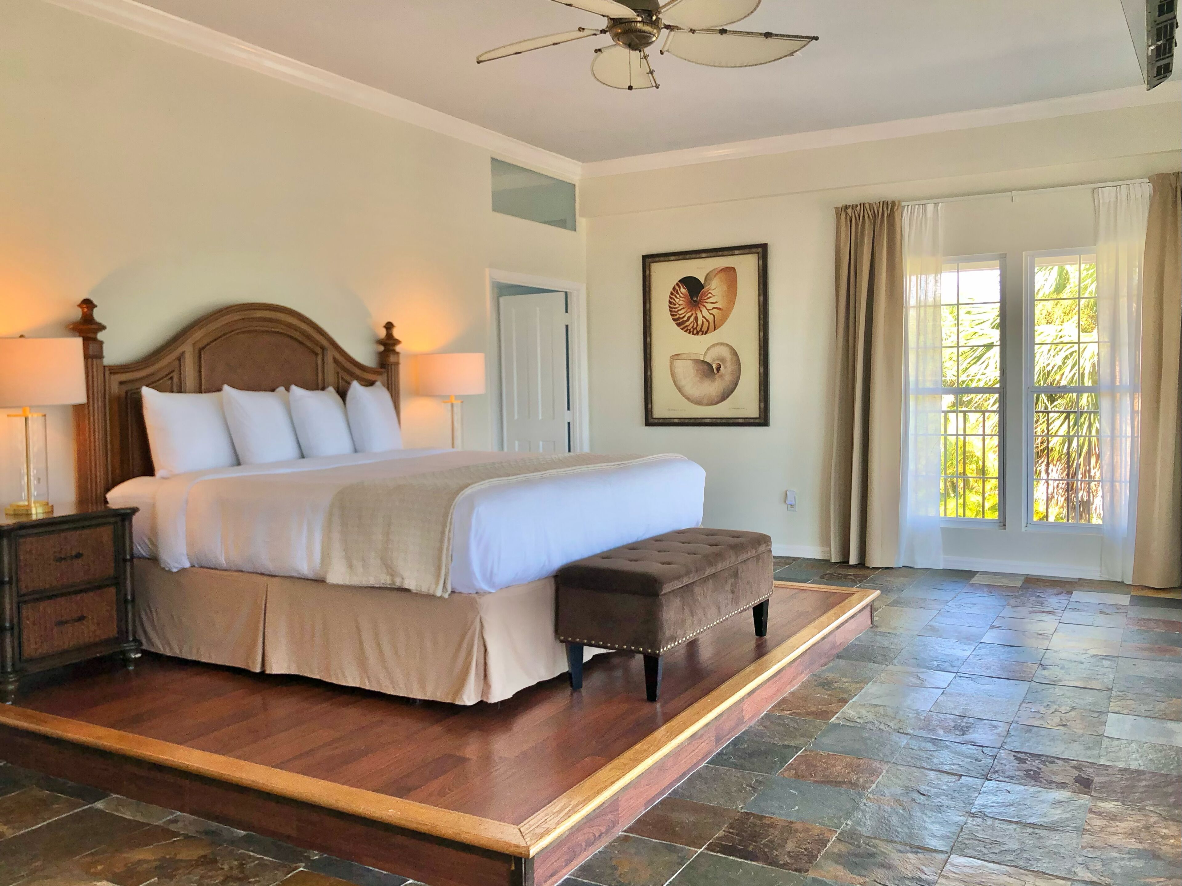 A spacious bedroom with a large bed, a wooden headboard, a nightstand with a lamp, a bench, and a ceiling fan.