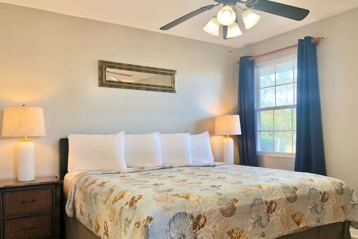 A bedroom with a bed, two bedside lamps, a ceiling fan, a window with curtains, and a mirror on the wall.