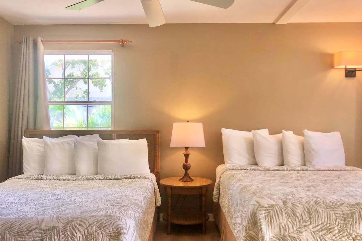 A hotel room with two beds, a ceiling fan, a window with curtains, a bedside lamp, and a wall-mounted light fixture.