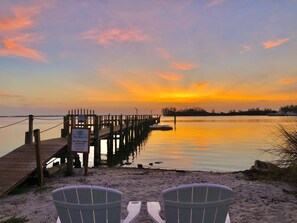 A wooden pier extending into calm waters with a sunset in the background. Two Adirondack chairs are placed on the sandy beach in the foreground. - Davis House Inn (Sebastian)