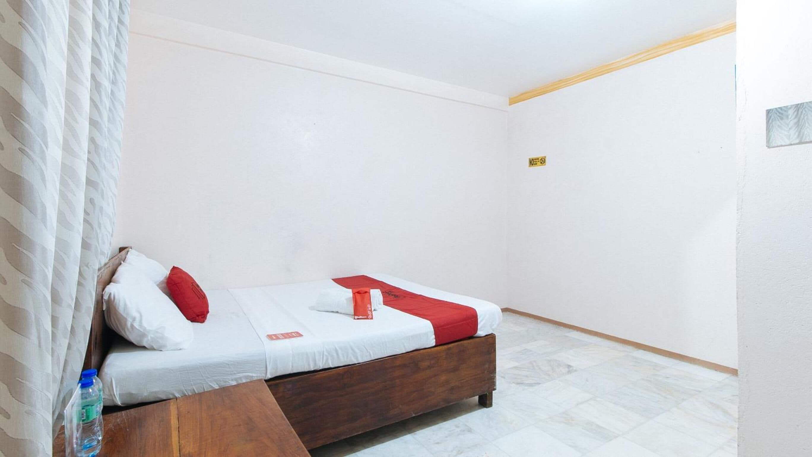Una habitación pequeña con cama individual, cabecera de madera, ropa de cama blanca y un cojín rojo decorativo.