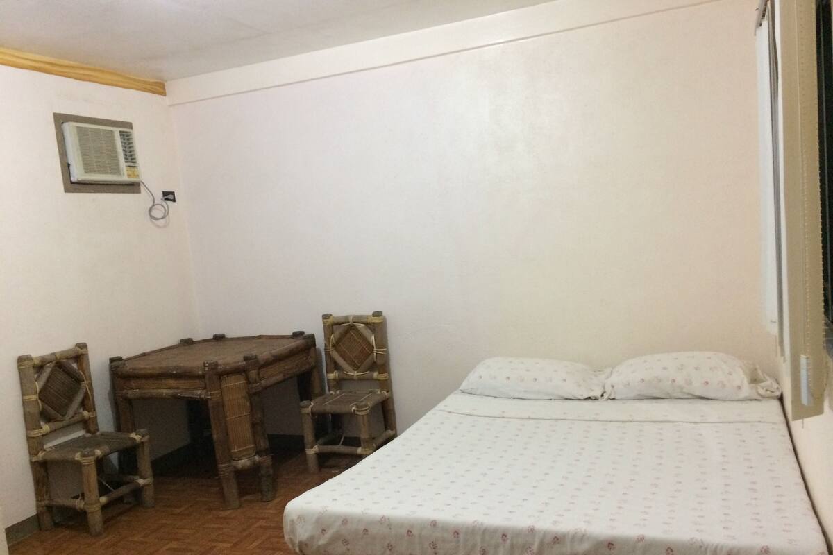 A small, simple room with a bed, a wooden table, and two chairs.