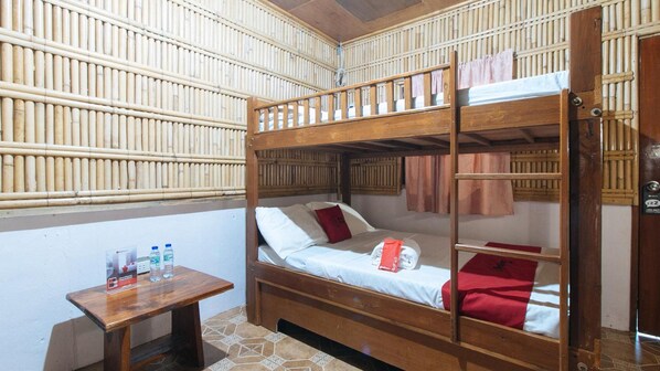 Quarto com beliches, paredes de bambu, uma pequena mesa de madeira com garrafas de água e uma toalha vermelha pendurada nos beliches.