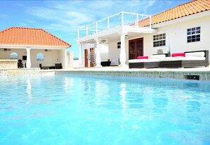 Ein Schwimmbecken mit überdachtem Bereich, ein weißes Gebäude mit roten Ziegeldach und ein Balkon mit Gartenmöbeln.