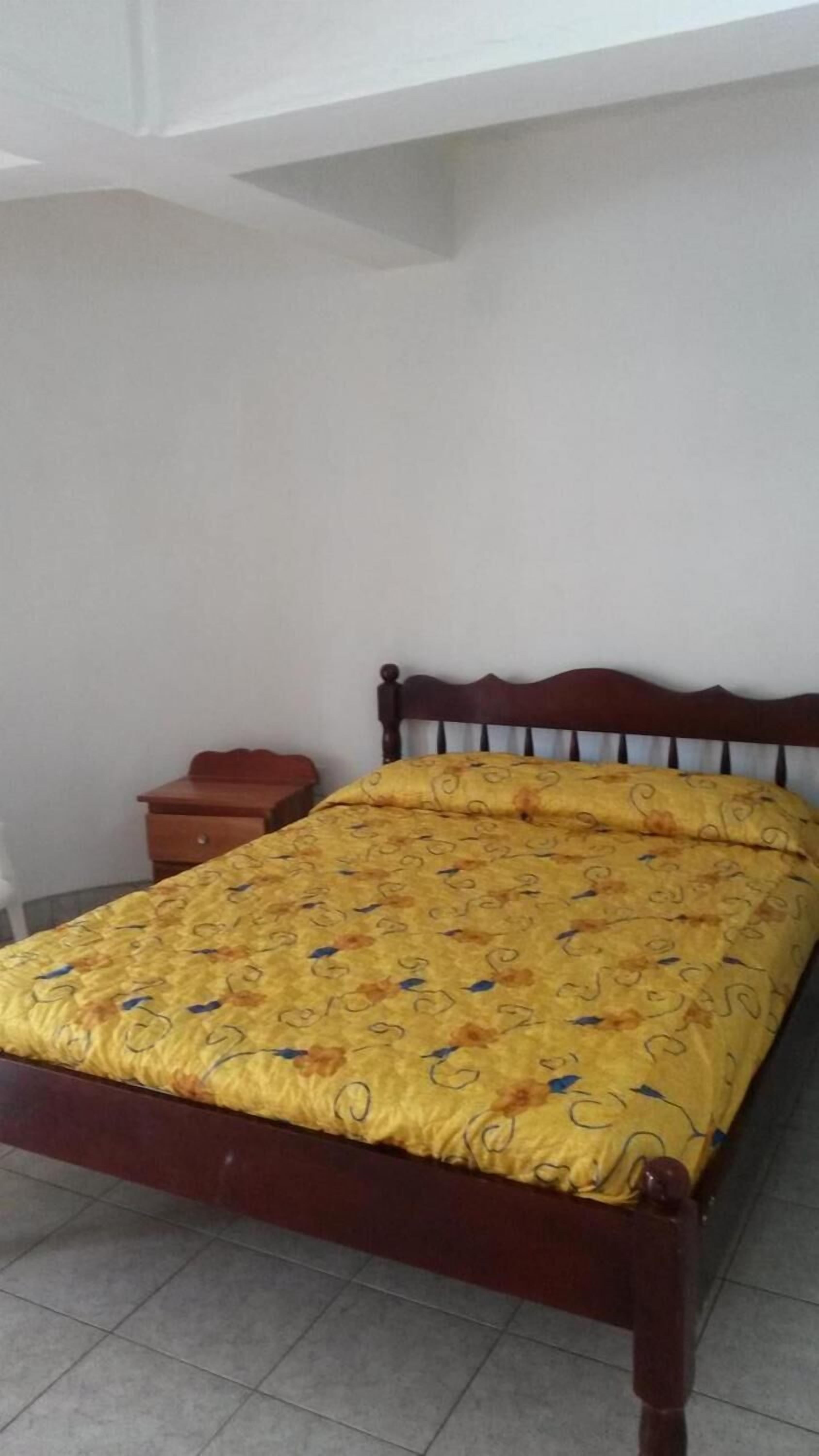 Una cama individual con estructura de madera y edredón amarillo estampado en una habitación con paredes blancas y suelo de baldosas.