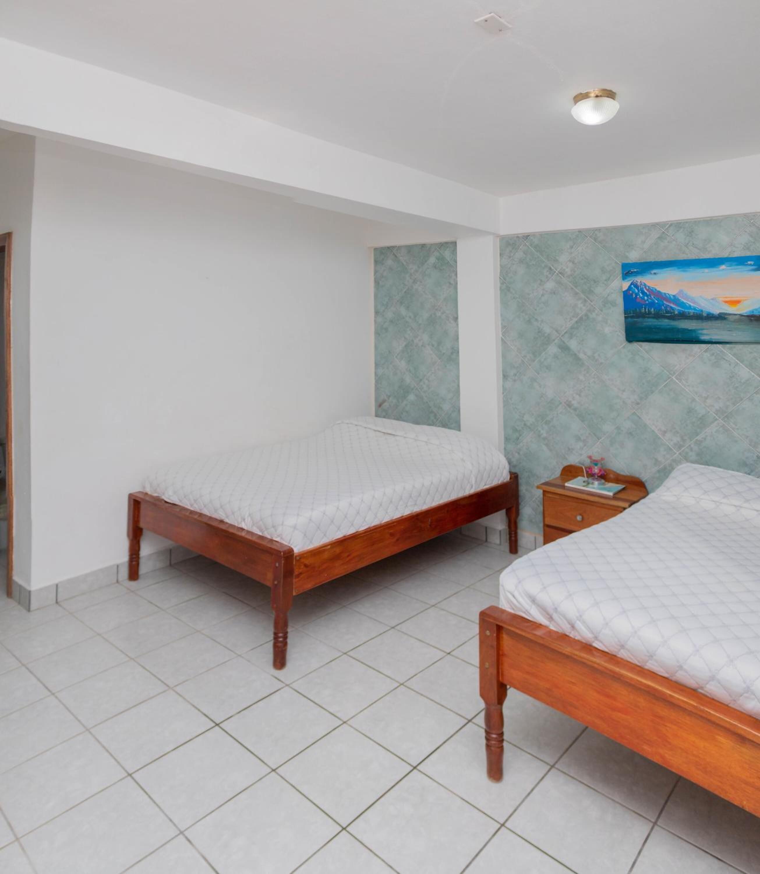 Ein Schlafzimmer mit zwei hölzernen Betten, einem kleinen Nachttisch, einem an der Wand hängenden Bild und einem Fliesenboden.