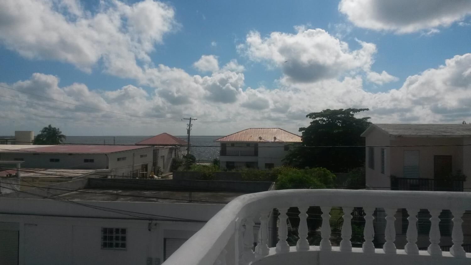 Zona residencial con edificios, un balcón y vista al mar bajo un cielo parcialmente nublado.