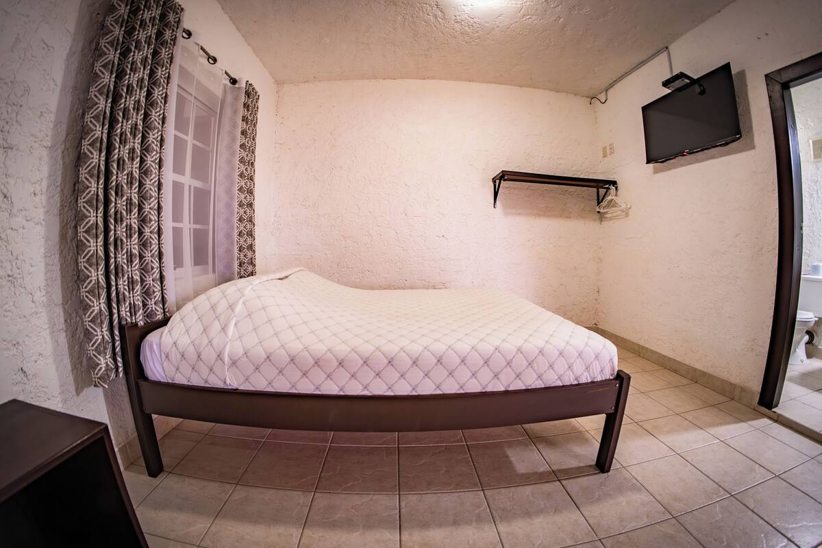 Une chambre à coucher avec un lit, une fenêtre ornée de rideaux à motifs, un téléviseur fixé au mur et un sol carrelé.