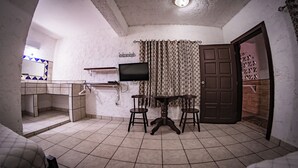 A compact kitchen with a tiled floor, a dining table with stools, a flat-screen TV mounted above the counter, and a patterned curtain. - Las Palmas Hotel (Corozal)