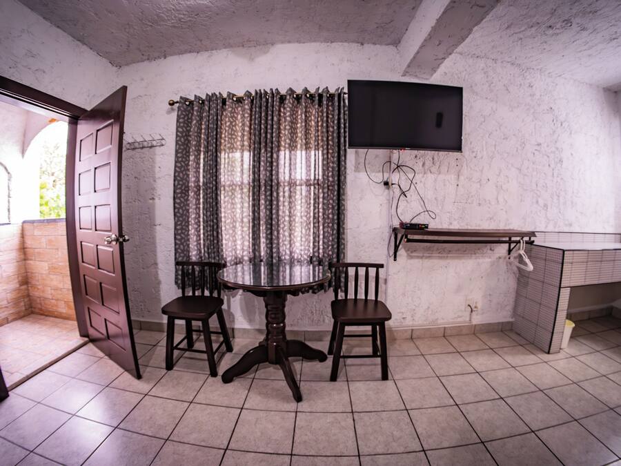 Una habitación con una mesa de comedor, dos sillas, un televisor montado en la pared y suelo de baldosas.