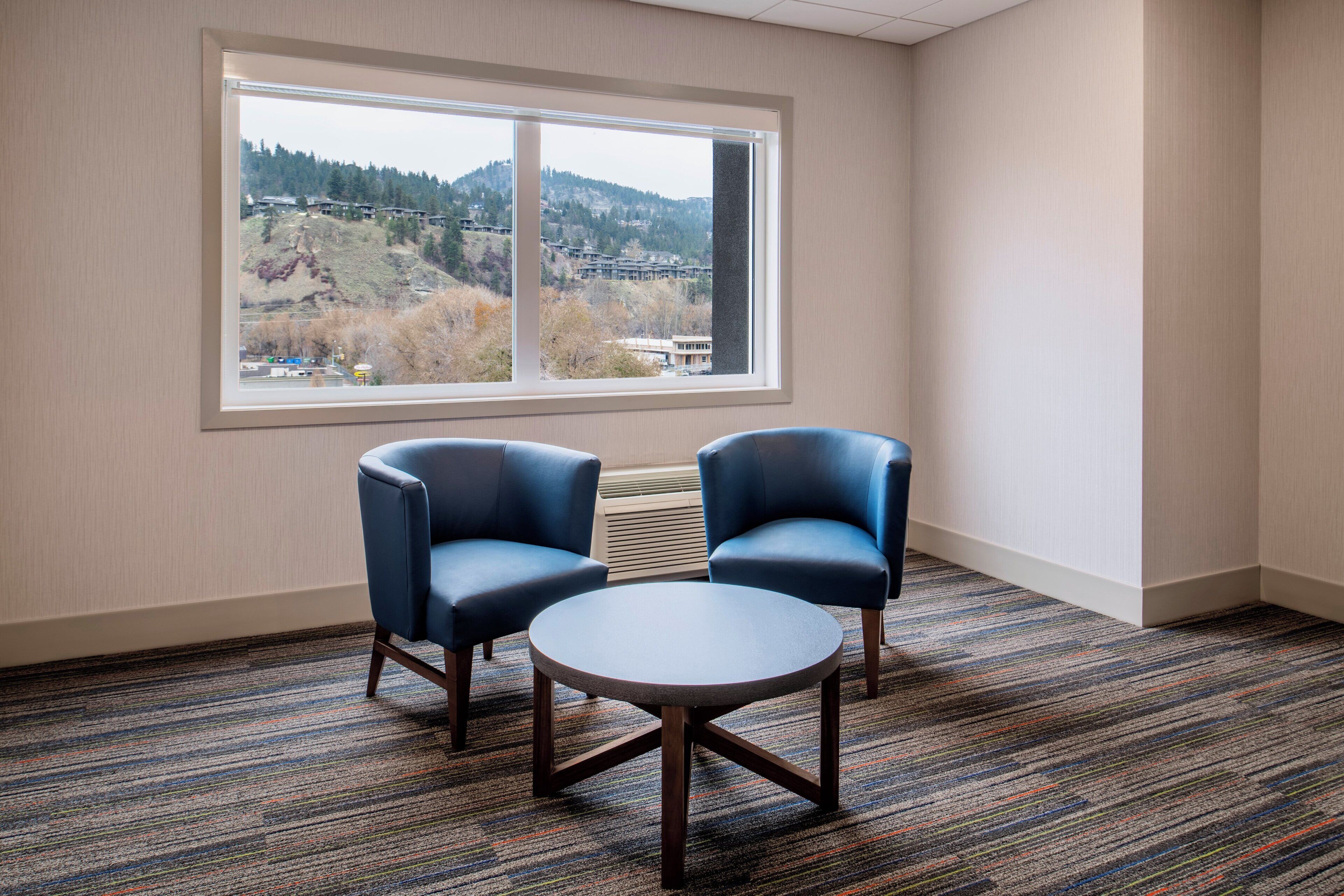 A room with two blue armchairs, a small round table, and a large window offering a view of a hilly landscape.