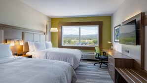 Ein Hotelzimmer mit zwei Betten, einem Schreibtisch, einem Sessel, einem Fernseher und einem großen Fenster mit Blick ins Freie.
