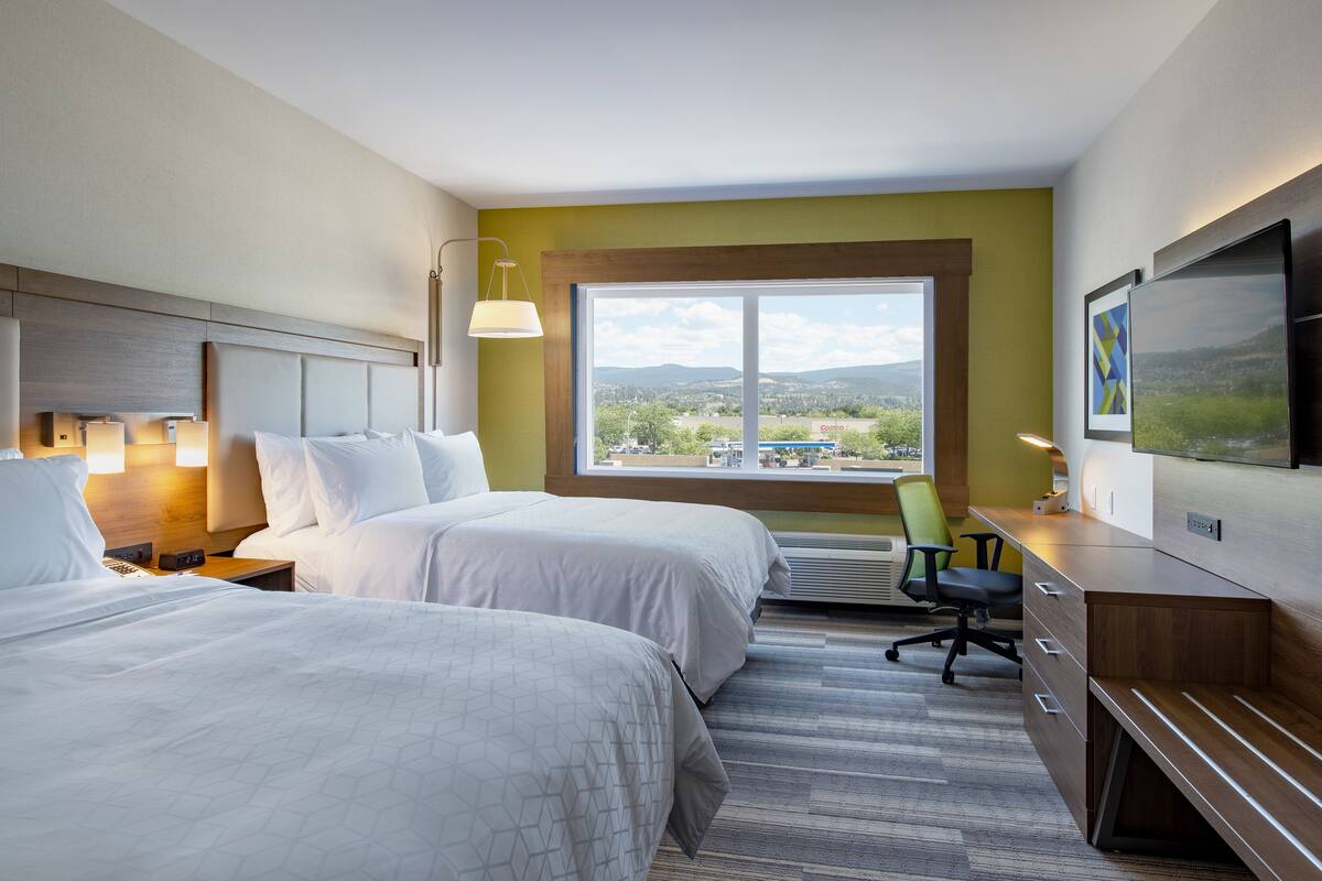 A hotel room with two beds, a desk, a chair, a TV, and a large window offering a view of the outdoors.