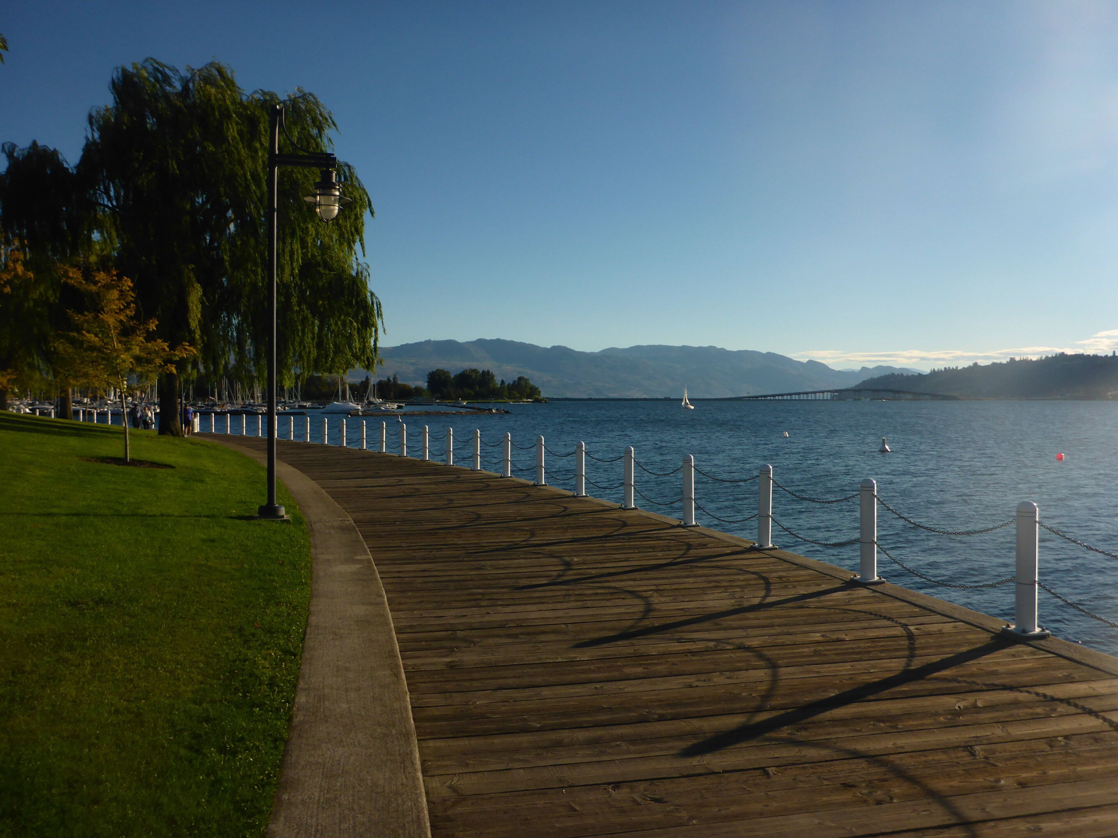 A wooden boardwalk along a waterfront with a clear view of the water and distant mountains.