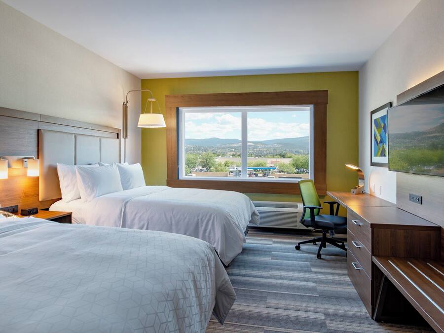 A hotel room with two beds, a desk, a chair, a TV, and a window with a view of a cityscape.