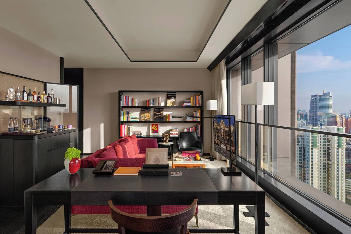 Uma sala de estar moderna com um sofá vermelho, uma mesa de centro, uma estante de livros e vista para a cidade através de grandes janelas.