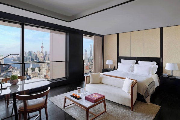 A modern hotel room with a large window offering a city skyline view, a neatly made bed, a small table with a book and fruit, and a sofa. - Bulgari Hotel Shanghai (Shanghai)