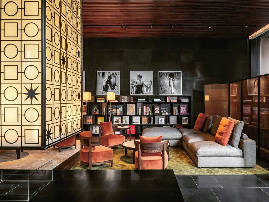 A modern lounge area with a geometric patterned wall, a sofa, armchairs, a coffee table, and a bookshelf with framed pictures.