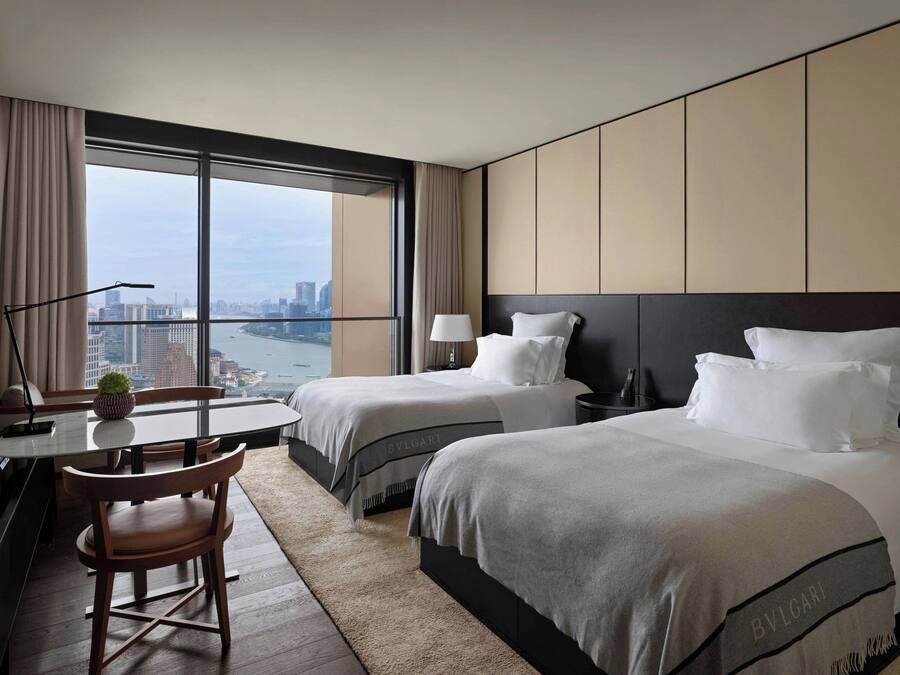 A hotel room with two beds, a dining table with chairs, a desk with a lamp, and a view of the city through large windows.