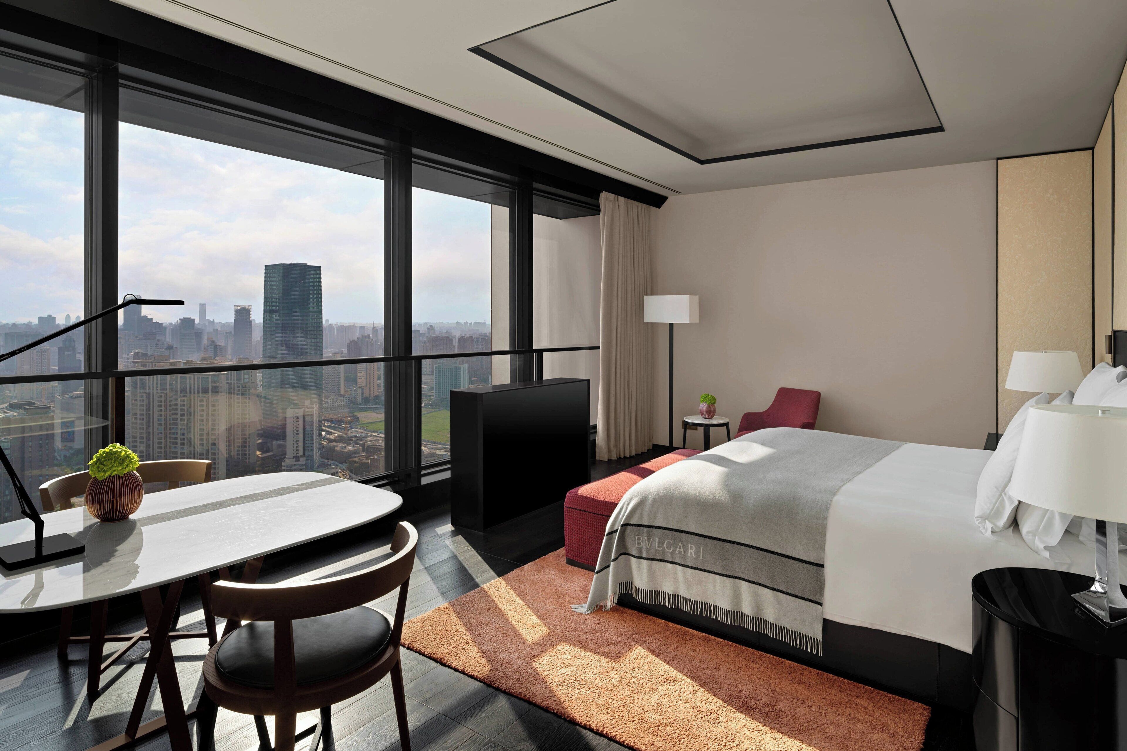 A modern hotel room with a large window offering a city view, a neatly made bed, a small table with chairs, and a desk with a lamp.