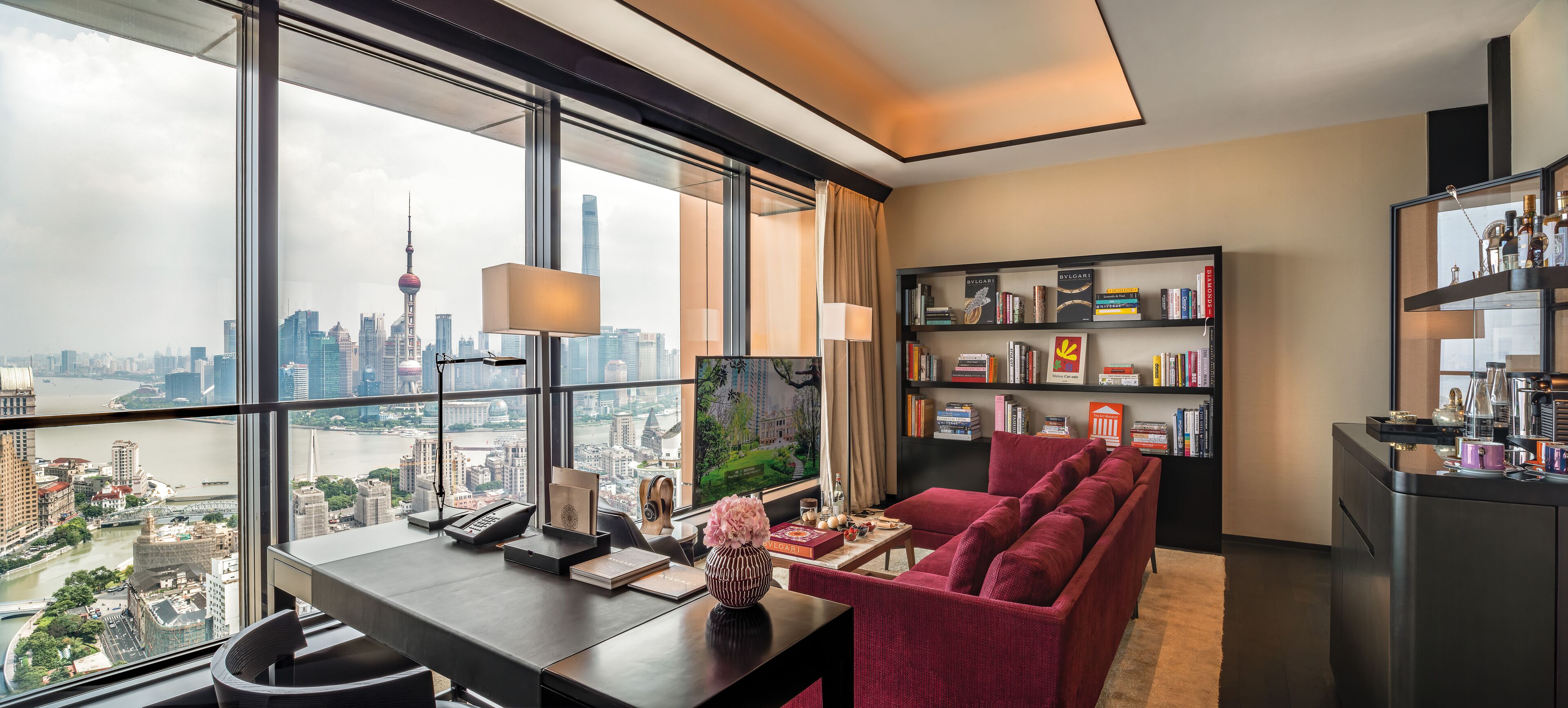 Uma sala de estar moderna com vista para a cidade, um sofá vermelho, uma estante de livros e uma mesa com luminária.