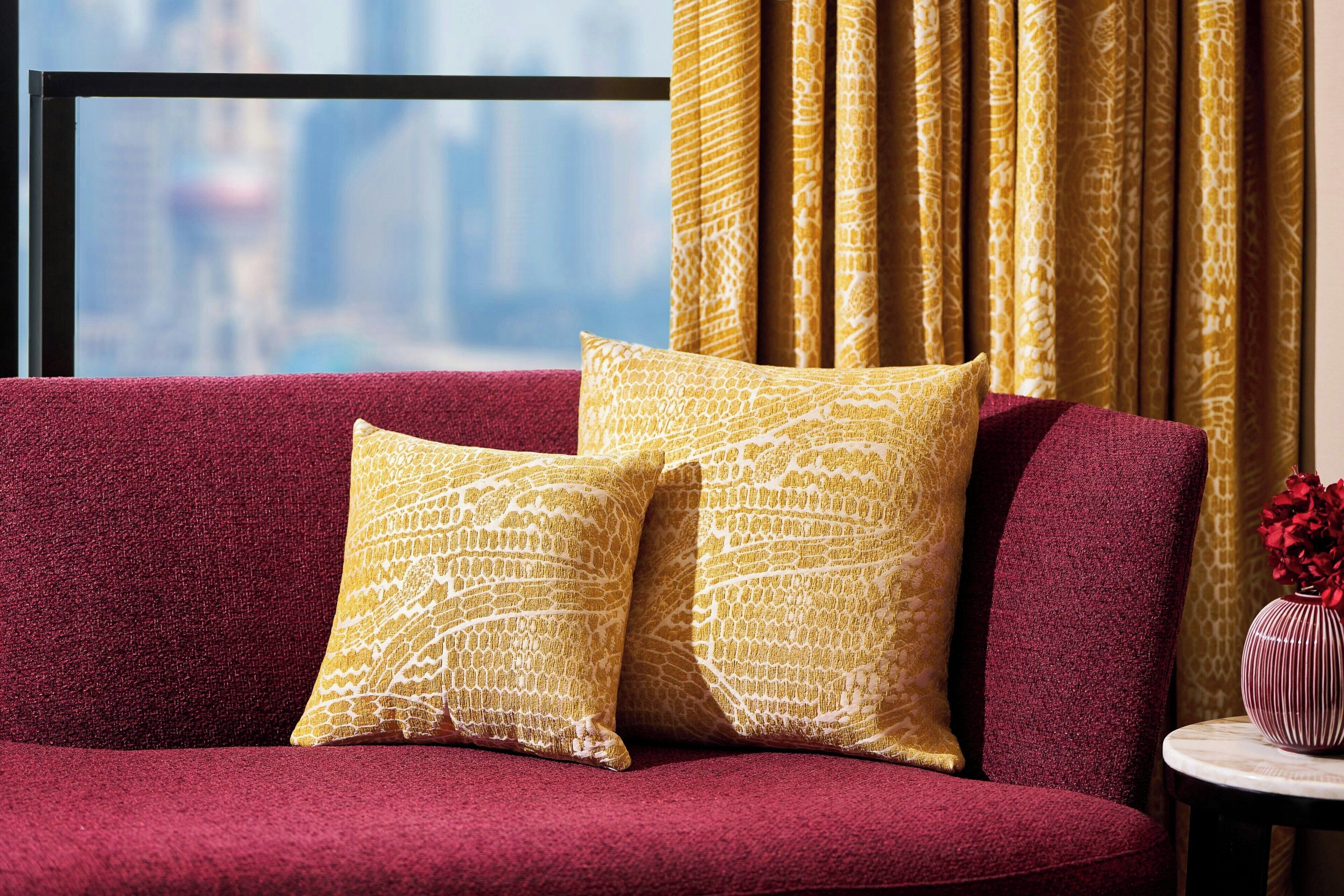 A red sofa with two yellow patterned cushions, a small round table with a vase of red flowers, and a view of a cityscape through a window with golden curtains.