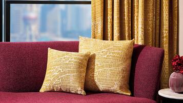 A red sofa with two yellow patterned cushions, a small round table with a vase of red flowers, and a view of a cityscape through a window with golden curtains.