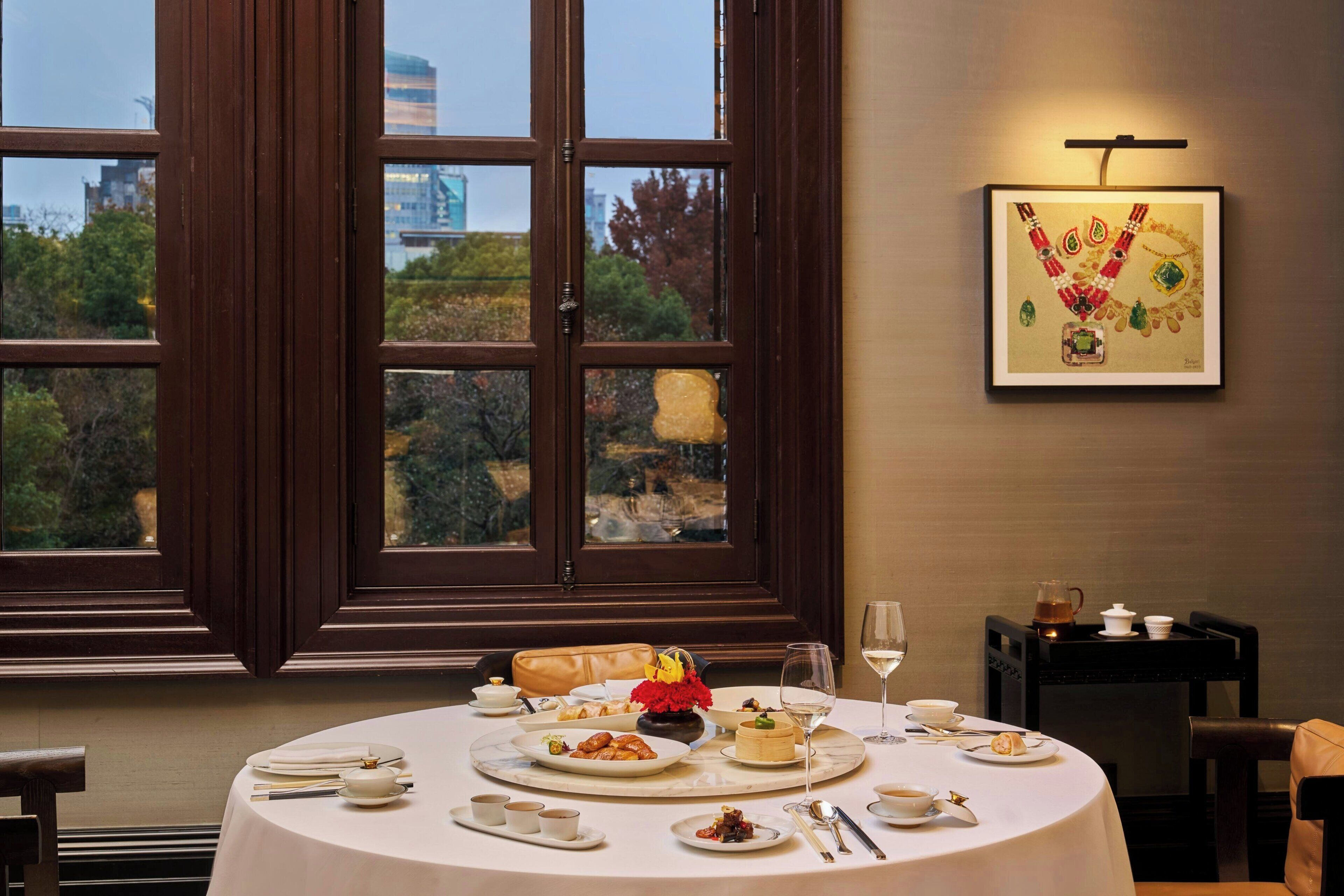 A dining area with a round table set for a meal, a framed artwork on the wall, and a view of the outdoors through large windows.