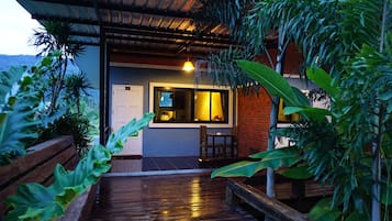 A wooden deck leading to a building with a glass door and a window, surrounded by lush greenery.