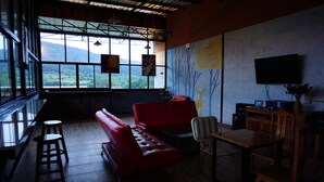 Una habitación con un sofá rojo, una mesa de madera y un televisor. Amplias ventanas ofrecen vistas al exterior con vegetación.